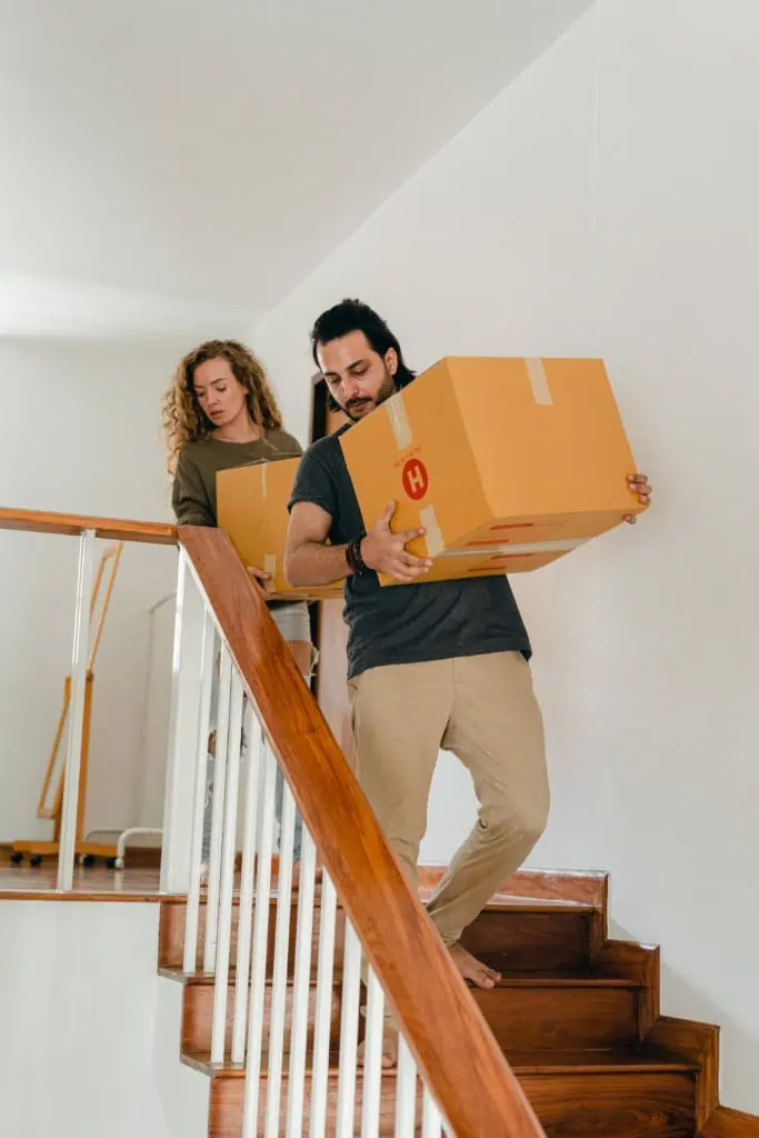 Two people carrying cardboard moving boxes down an interior staircase