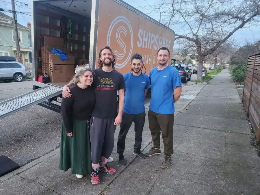 Three ShipShape crew members in blue shirts standing on the sidewalk with a customer beside the loaded truck at the end of a job.